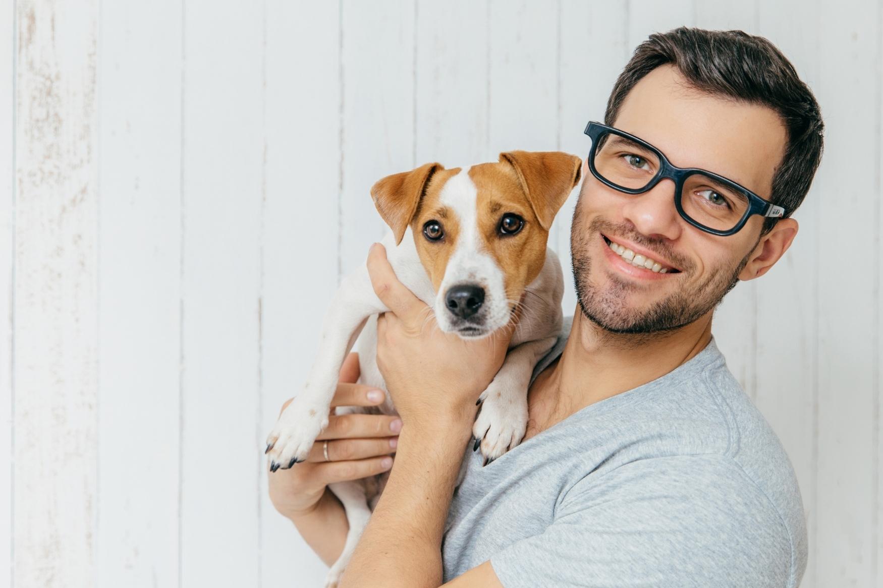 A man with glasses holding a small dog with brown and white markings against a white wooden background