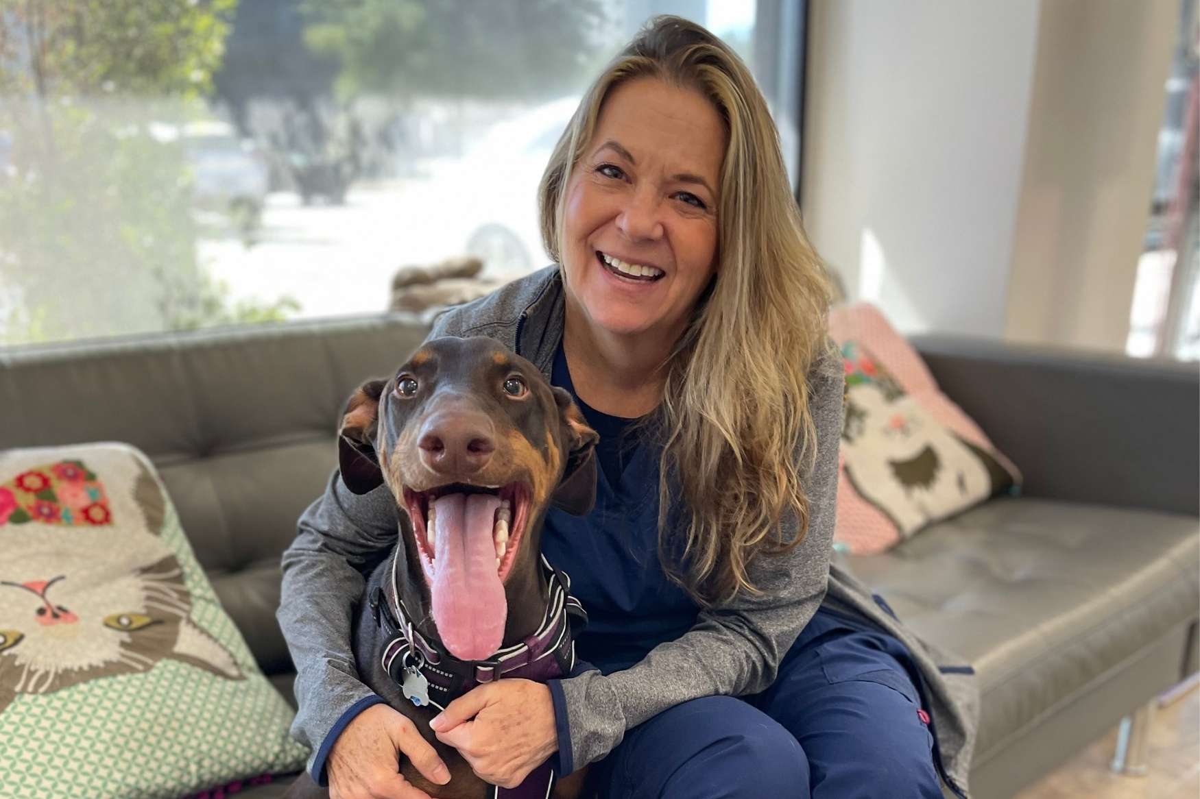A person in scrubs sits on a gray sofa with a happy Doberman Pinscher on their lap